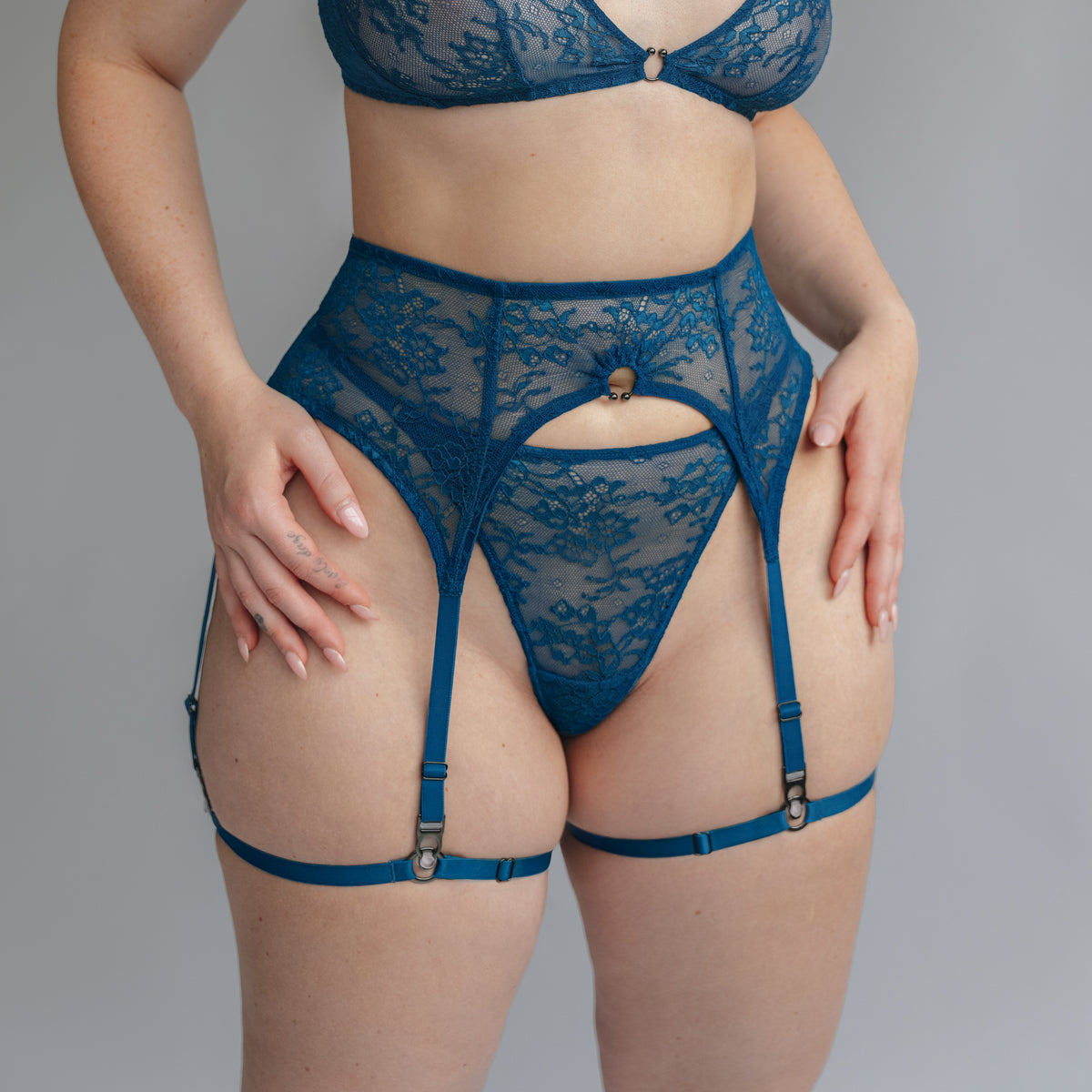 model wearing a teal blue lace bralette garter belt and thong with horseshoe ring details