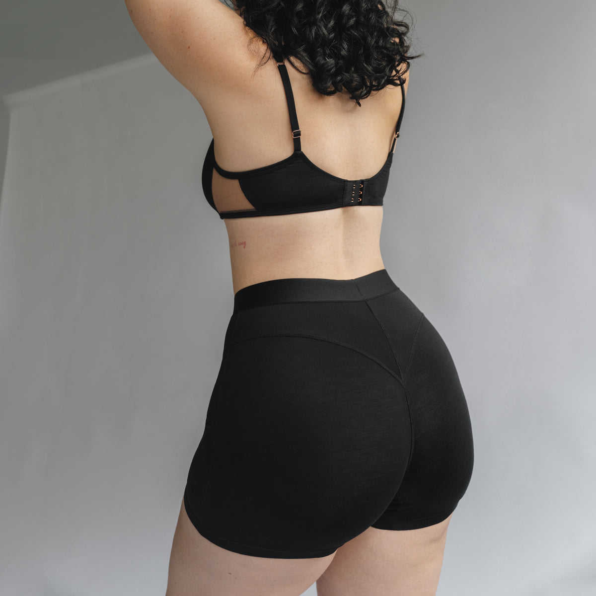 Back view of a model wearing a black bralette with back closure and a high waisted women boxer in black with front seaming detail and wide elastic band