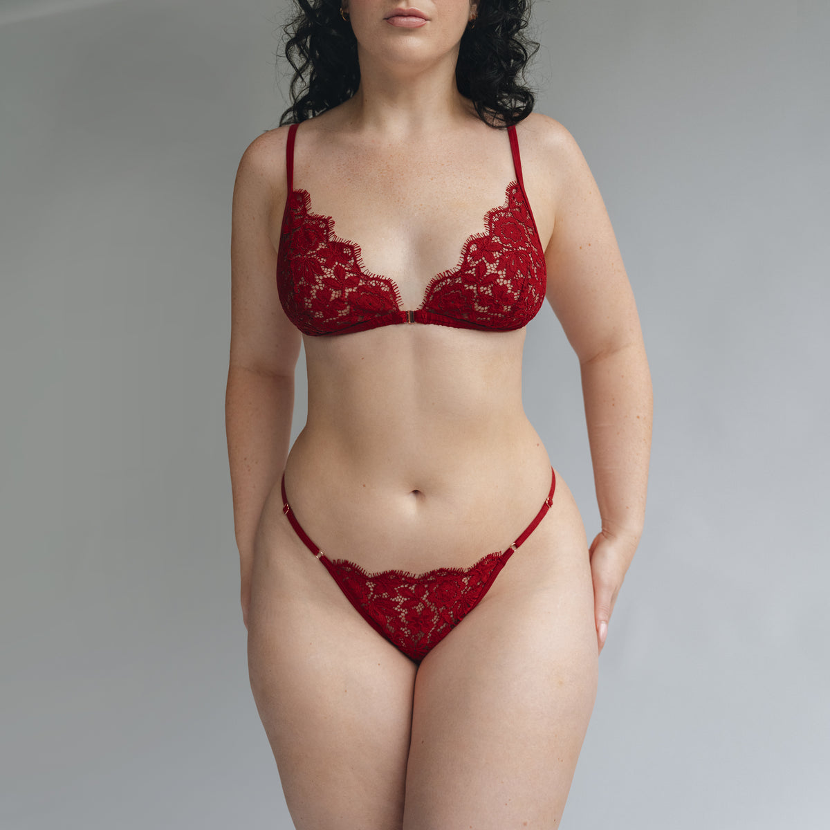 Model wearing a red lace triangle cup bralette and thong with adjustable waist elastics